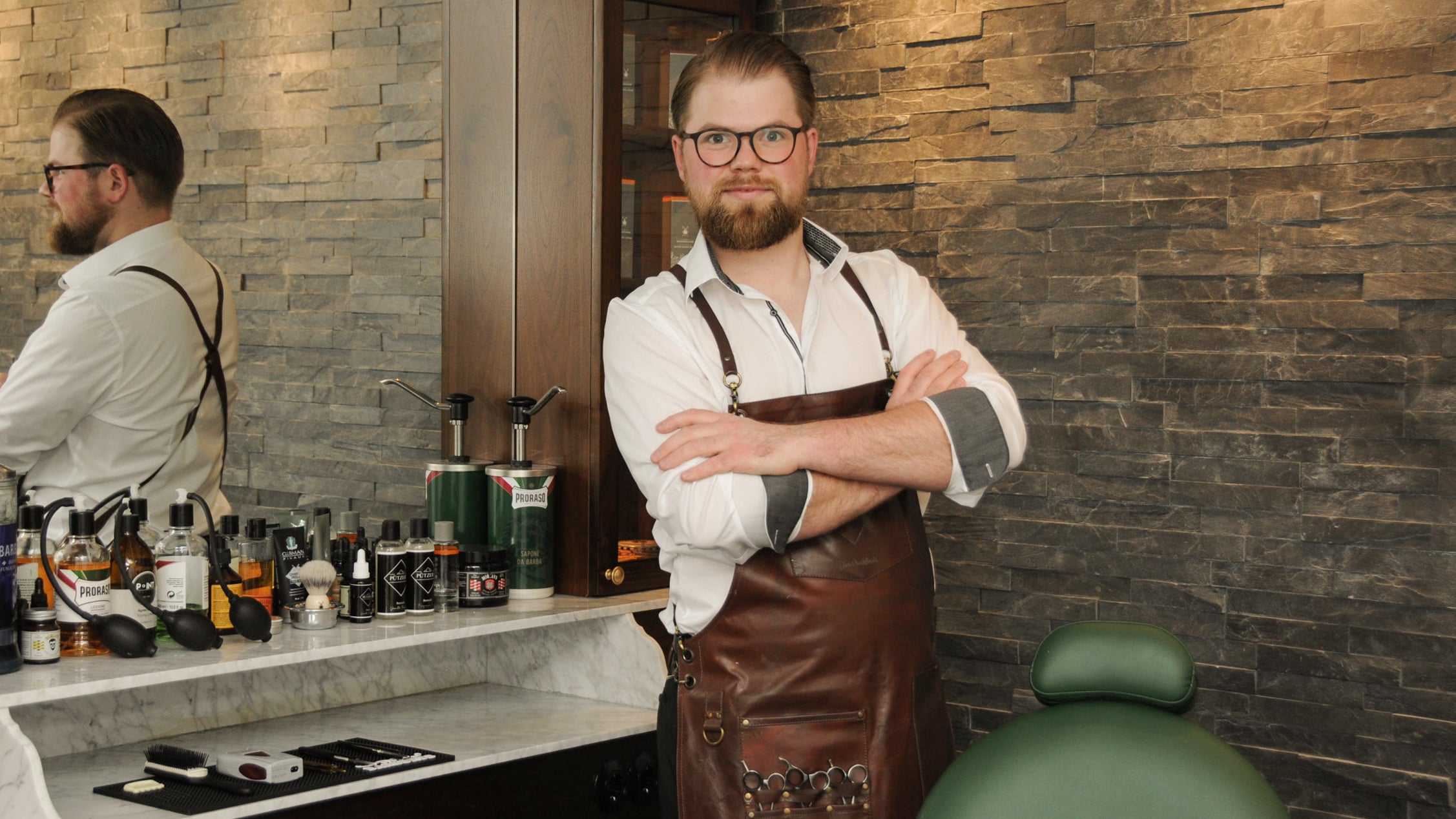 Barber in leather apron standing confidently in a stylish barbershop with grooming products on counter and a brick accent wall in the background.