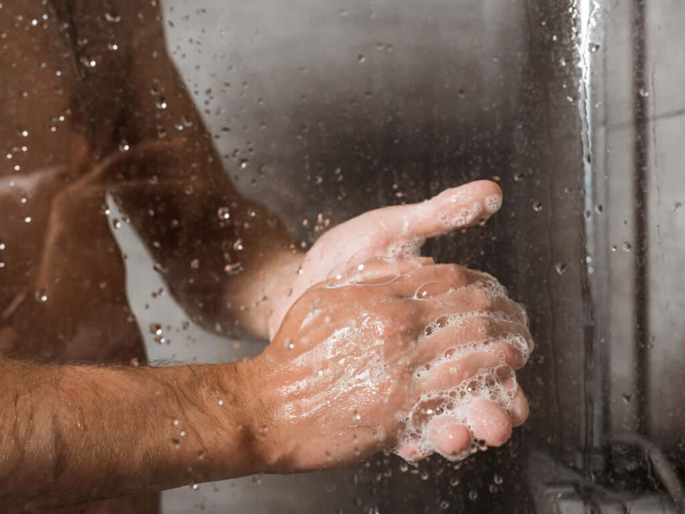 Nahaufnahme von zwei nassen Händen, die in einer Dusche mit Seife gewaschen werden, umgeben von Wassertropfen auf einer Glasscheibe. 